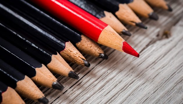 Red pencil stands out from the crowd of black  on a wooden white background.