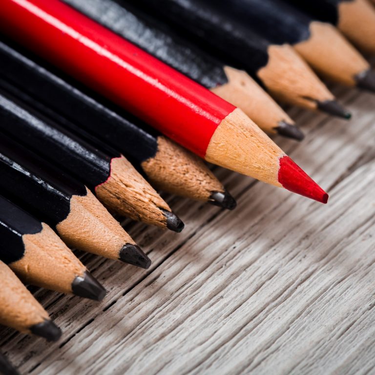 Red pencil stands out from the crowd of black  on a wooden white background.